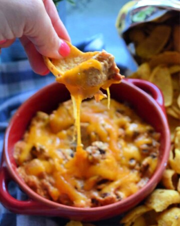 Manicured hand scooping up chili cheese dip from red bowl with frito lay chip.