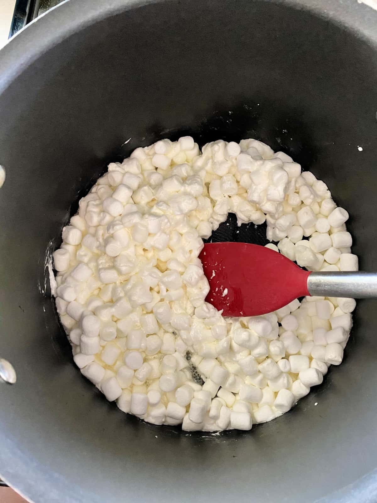 Marshmallows and butter melting in a large stock pot being stirred with red spatula.