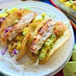 White round plate with three soft fish tacos with avocado and cabbage on them.