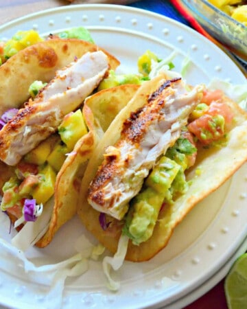 White round plate with three soft fish tacos with avocado and cabbage on them.