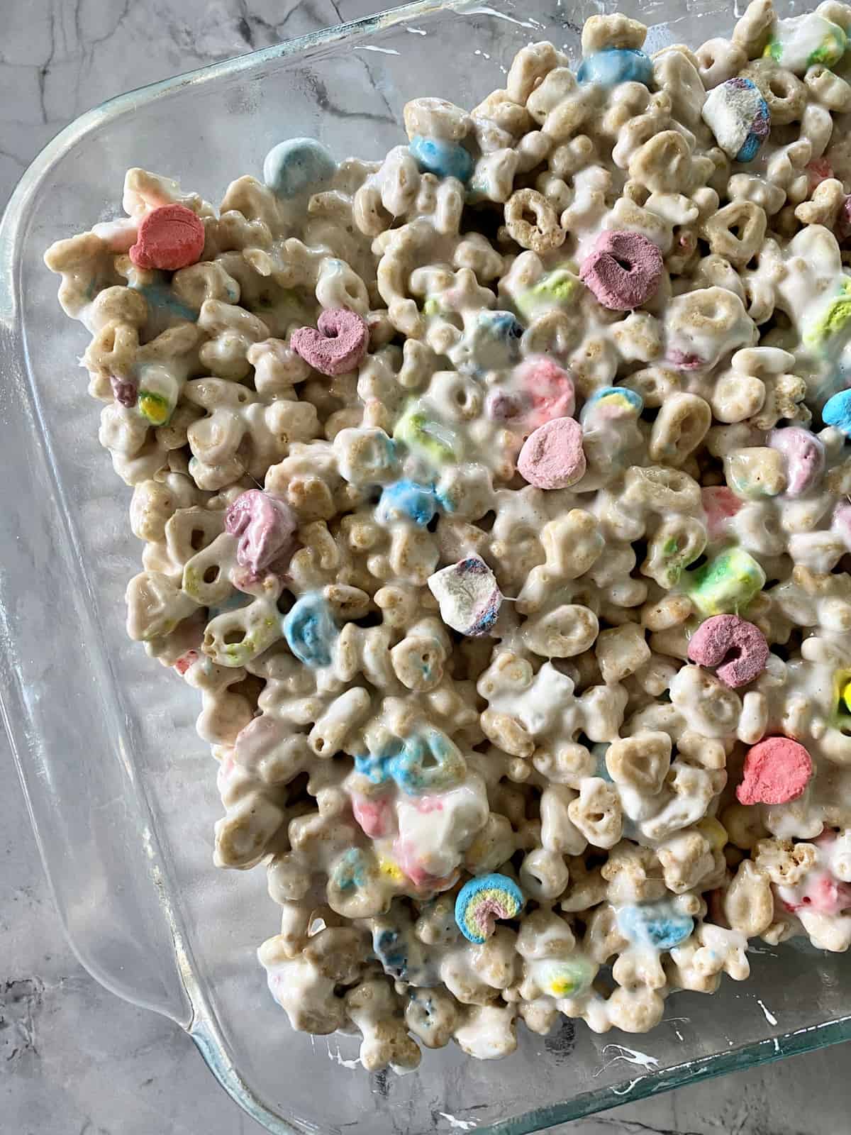 Closeup top view of lucky charms, melted marshmallows, and butter flattened into glass casserole dish.