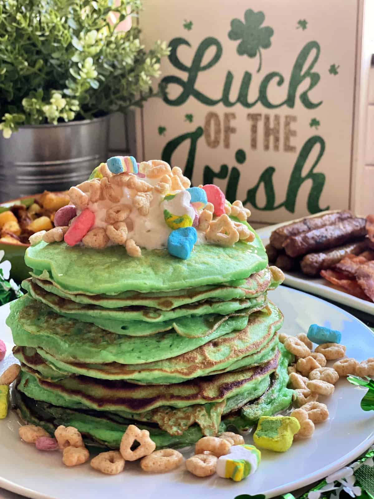 side view of plated stacked green pancakes topped with lucky charms with sausage in background.