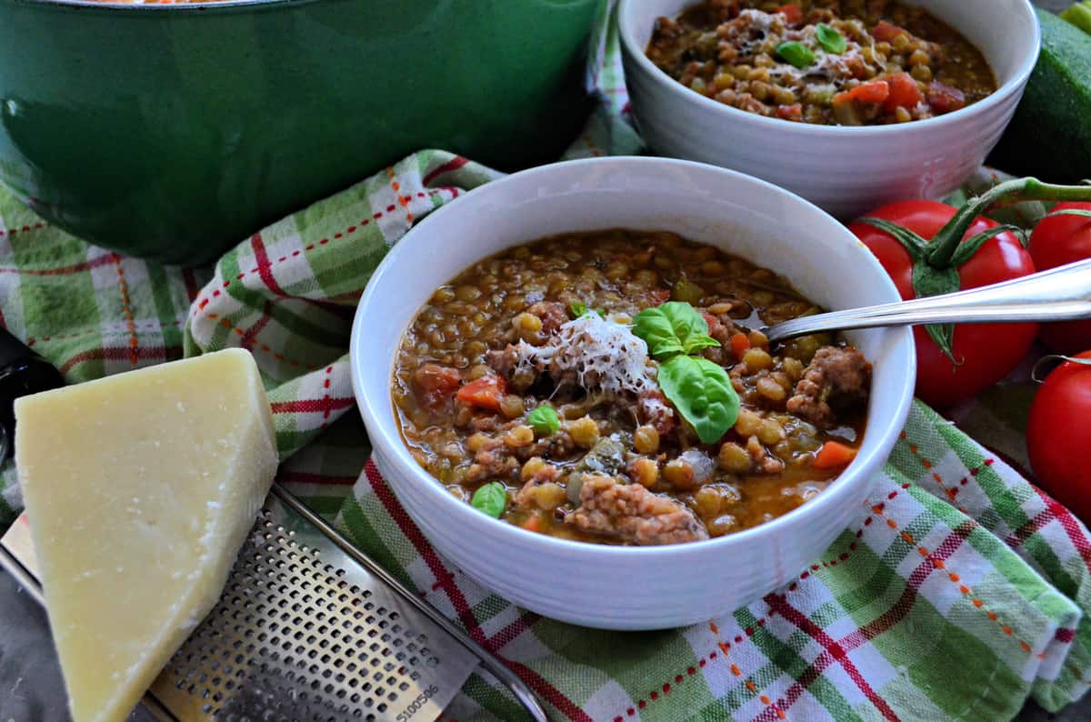 two white bowls filled with Sausage Lentil Soup next to fresh tomatoes, parmesan and grater on tablecloth.