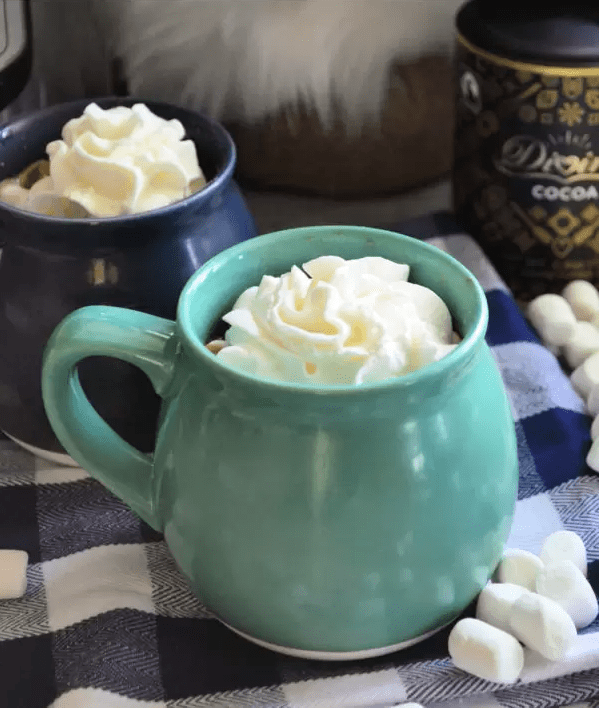 Two mugs with whipped cream inside on checkered tablecloth decoratively placed amongst mini marshmallows.