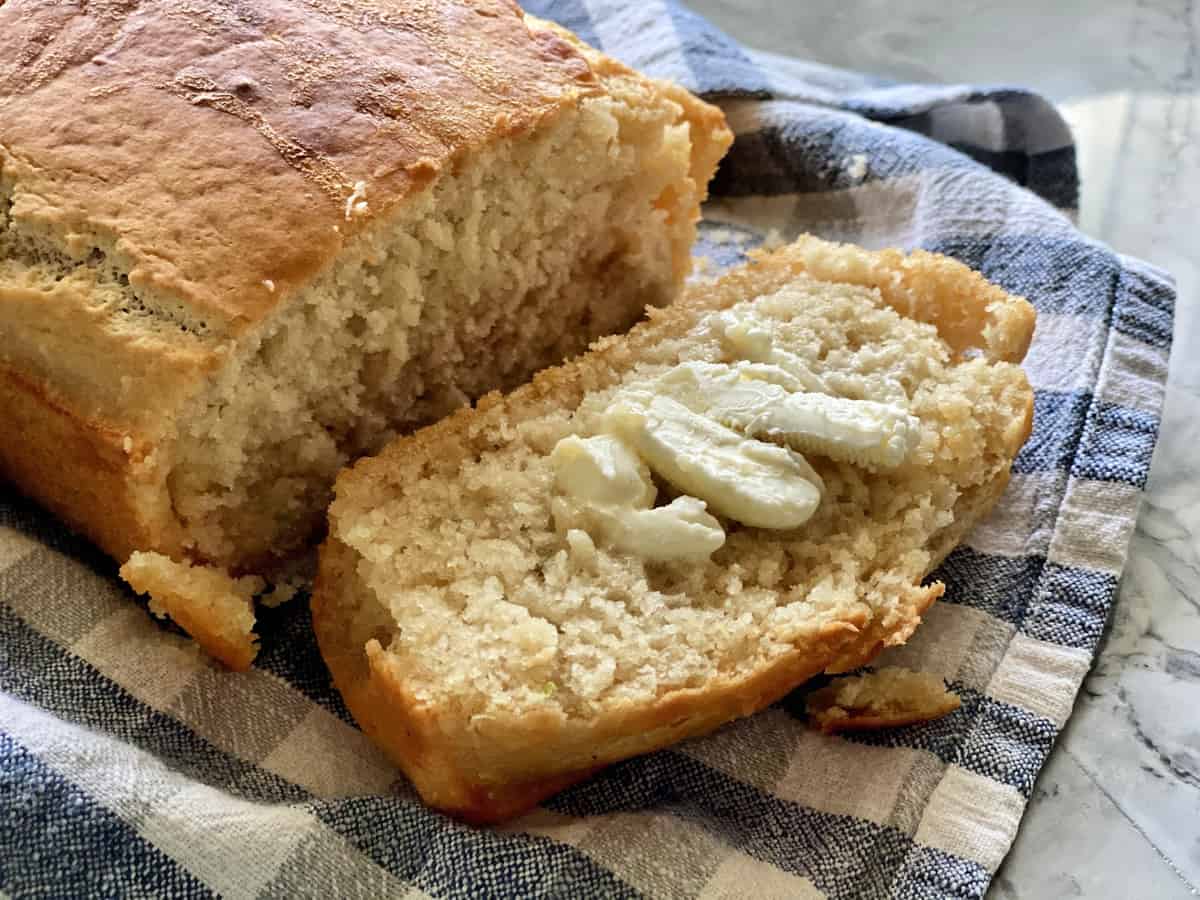 Loaf with piece of beer bread sliced off with butter on blue checkered tablecloth.