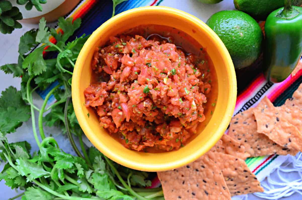 Top view of a bowl of fresh salsa with chips, cilantro, lime, and jalapeno around it.