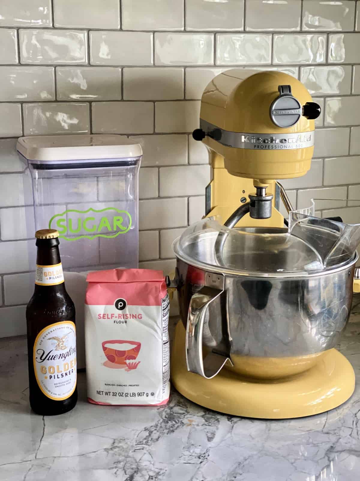 Yuengling Pilsner, self-rising Flour, and sugar on countertop next to yellow KitchenAid Professional 600 mixer.