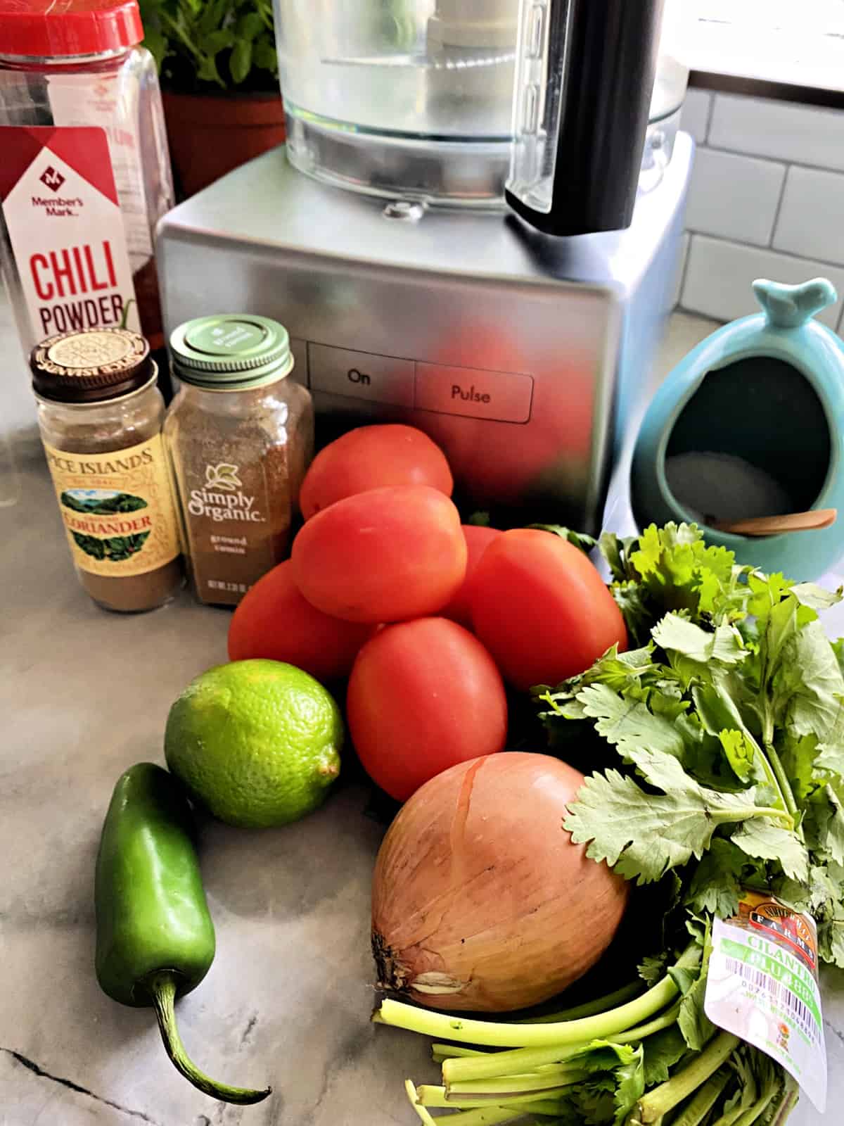 Ingredients on counter; spices, plum tomatoes, lime, jalapeno, onion, cilantro and food processor.