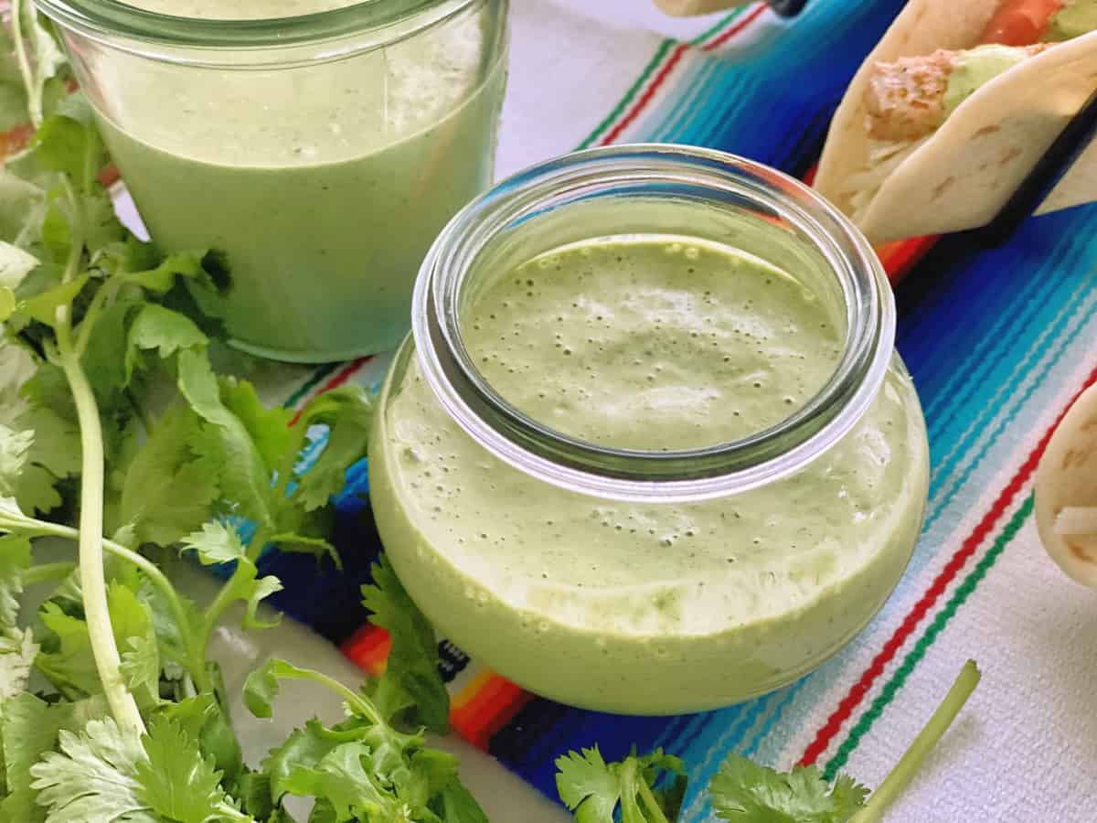 closeup jar of creamy light green liquid on colorful tablecloth with fresh cilantro and fish tacos.