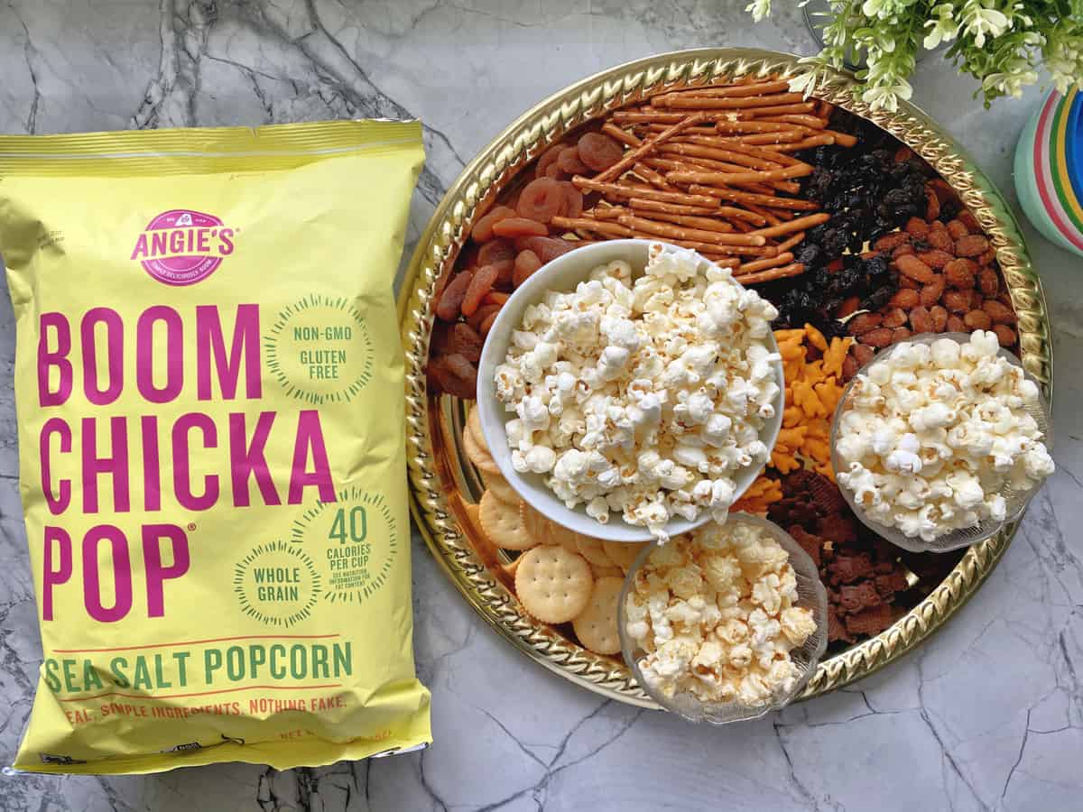 Angie’s BOOMCHICKAPOP Popcorn next to Snack Board of popcorn, dried fruit, pretzels, crackers, and nuts.