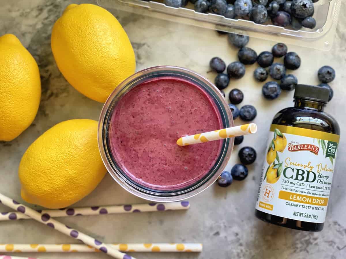 Top view Barlean's Seriously Delicious CBD Lemon Drop bottle next to smoothie, fresh blueberries, and lemons.