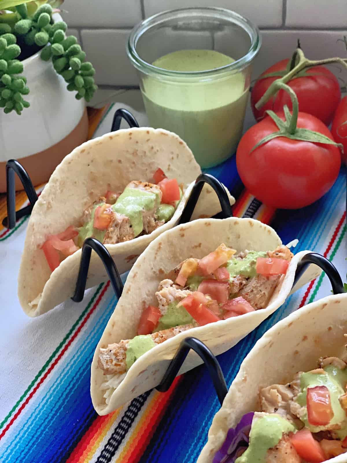 3 Blackened Swordfish Tacos in wire holder with glass of cilantro dressing and tomatoes in background.