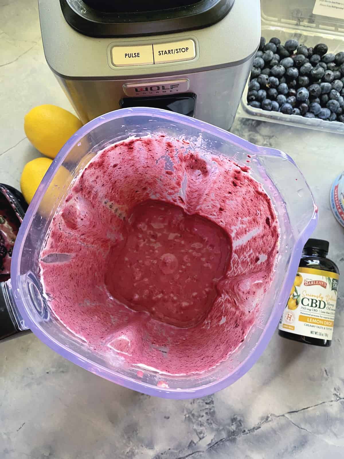 Top view of blender with purple blended liquid next to CBD oil bottle, blueberries, and lemons.