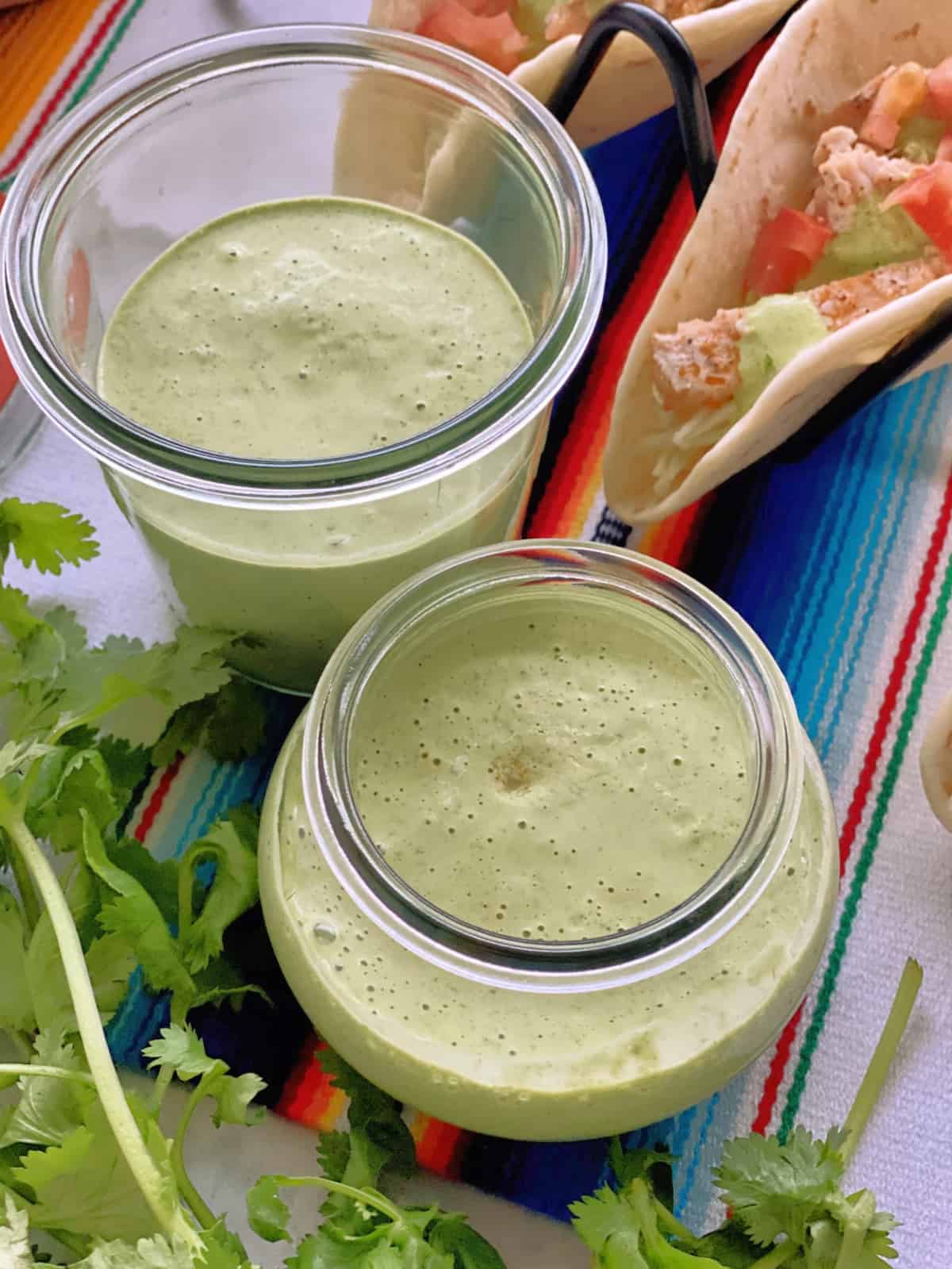 two jars of creamy light green liquid on colorful tablecloth with fresh cilantro and fish tacos.