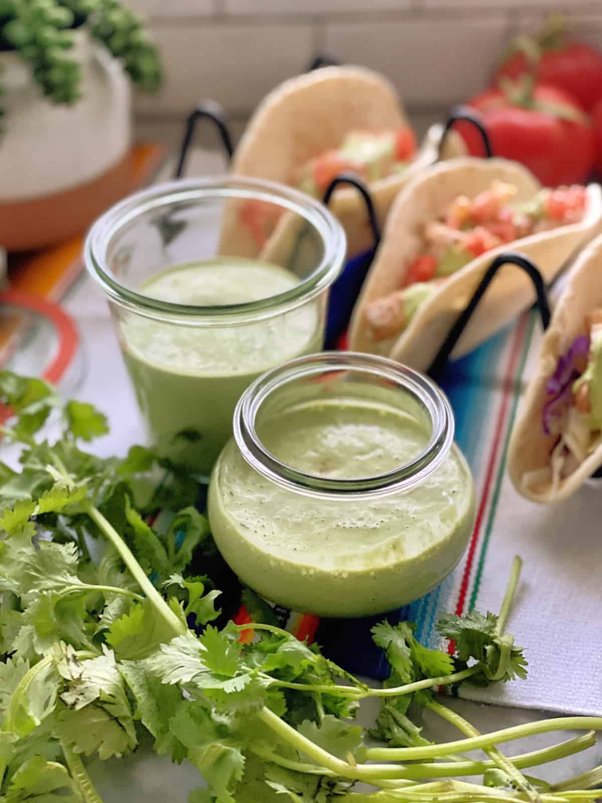 two jars creamy cilantro dressing on colorful tablecloth with fresh cilantro near and fish tacos behind.