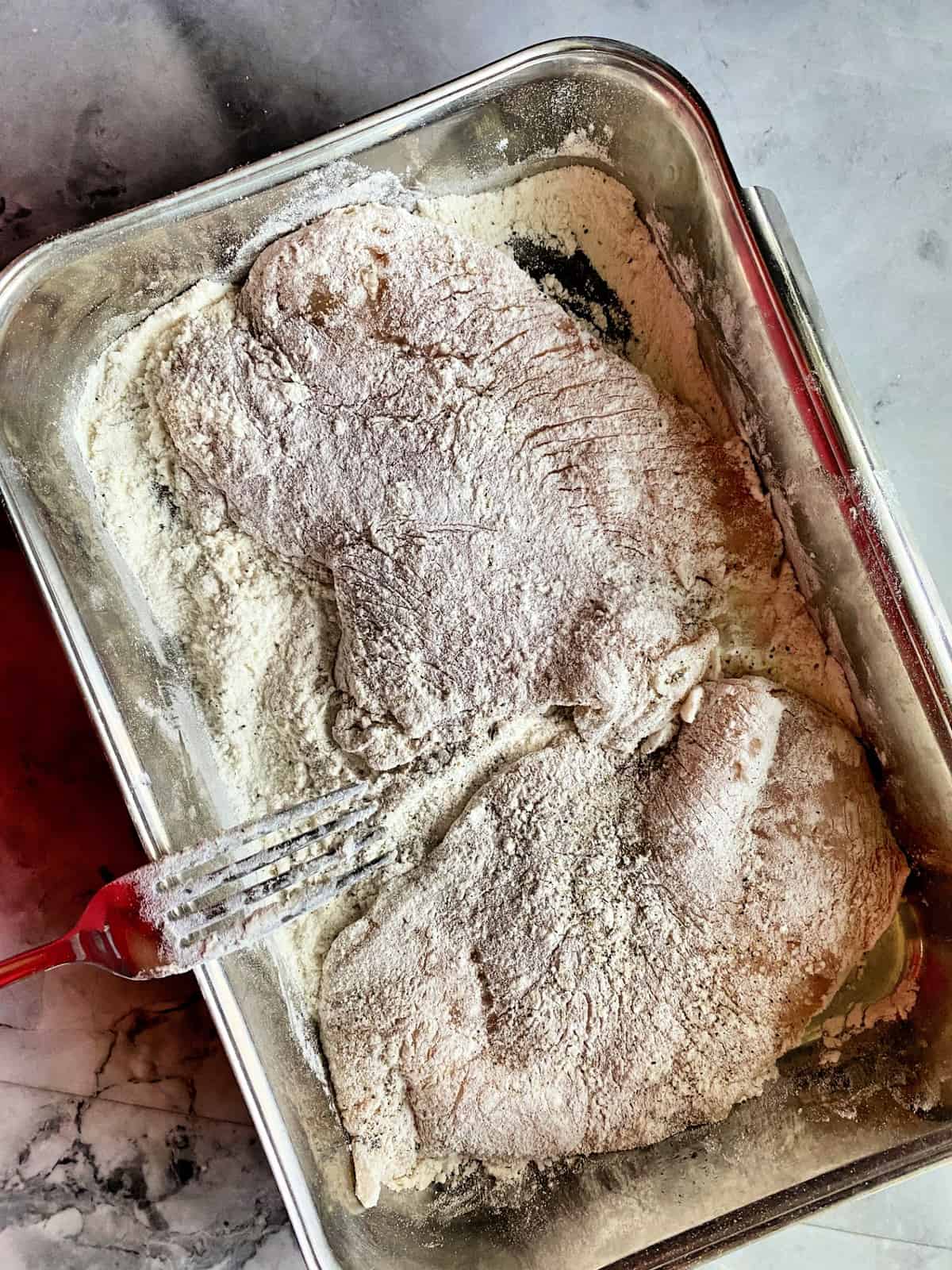 Breaded chicken cutlets in tin baking pan with fork and breading flower.