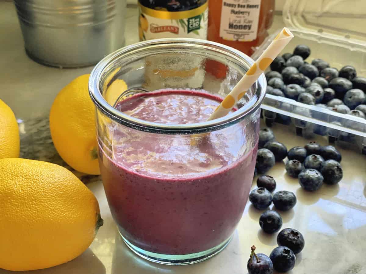Side view of purple smoothie in glass with paper straw on counter with lemons and fresh blueberries.
