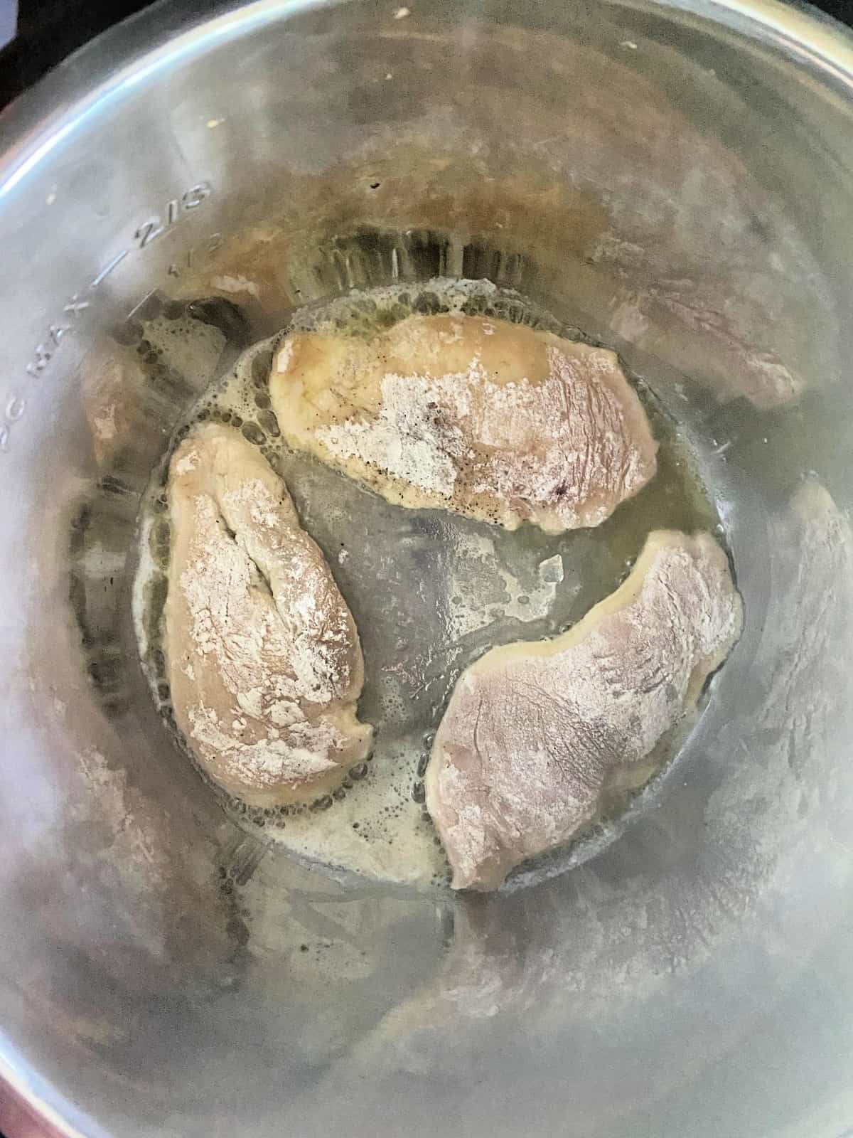 3 Chicken cutlets cooking in the Instant Pot covered in flower with thin liquid layer bubbling at bottom.