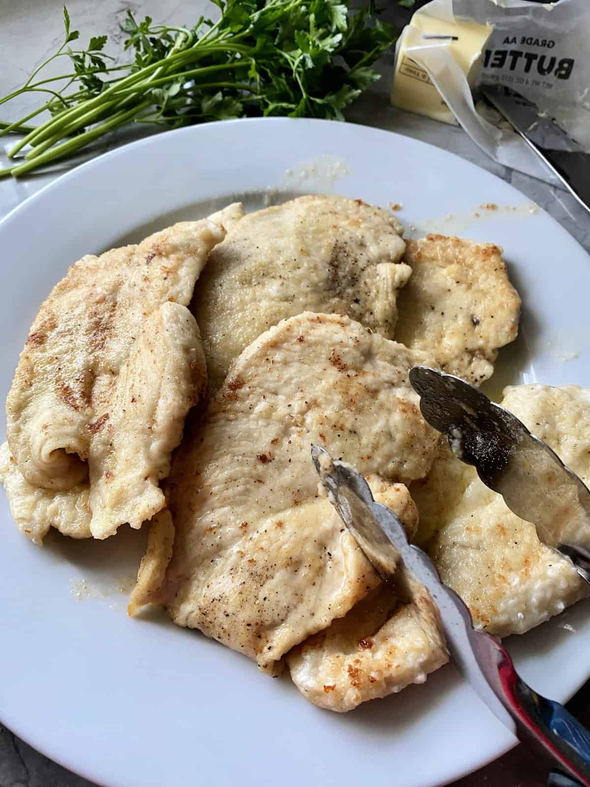 5 Browned chicken cutlets on a white plate with metal tongs.