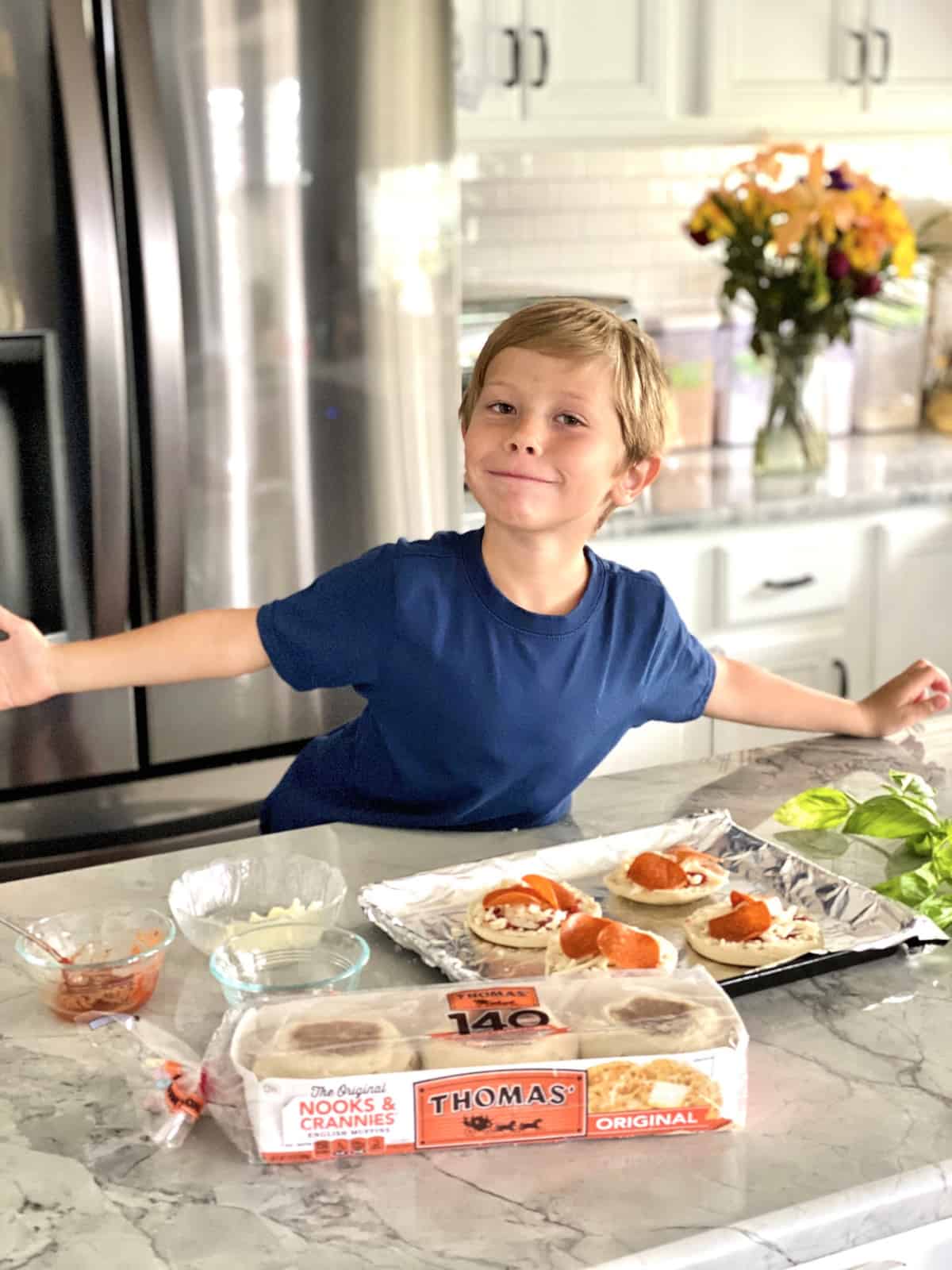 Little Boy making Toaster Oven English Muffin Pizzas on aluminum foil covered pan with ingredients displayed.