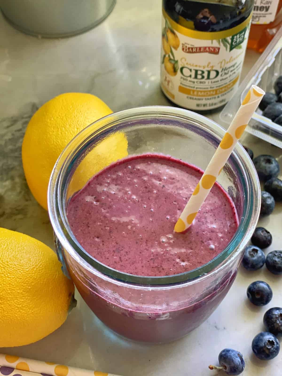Top view of CBD purple smoothie in glass with paper straw next to blueberries and lemons.