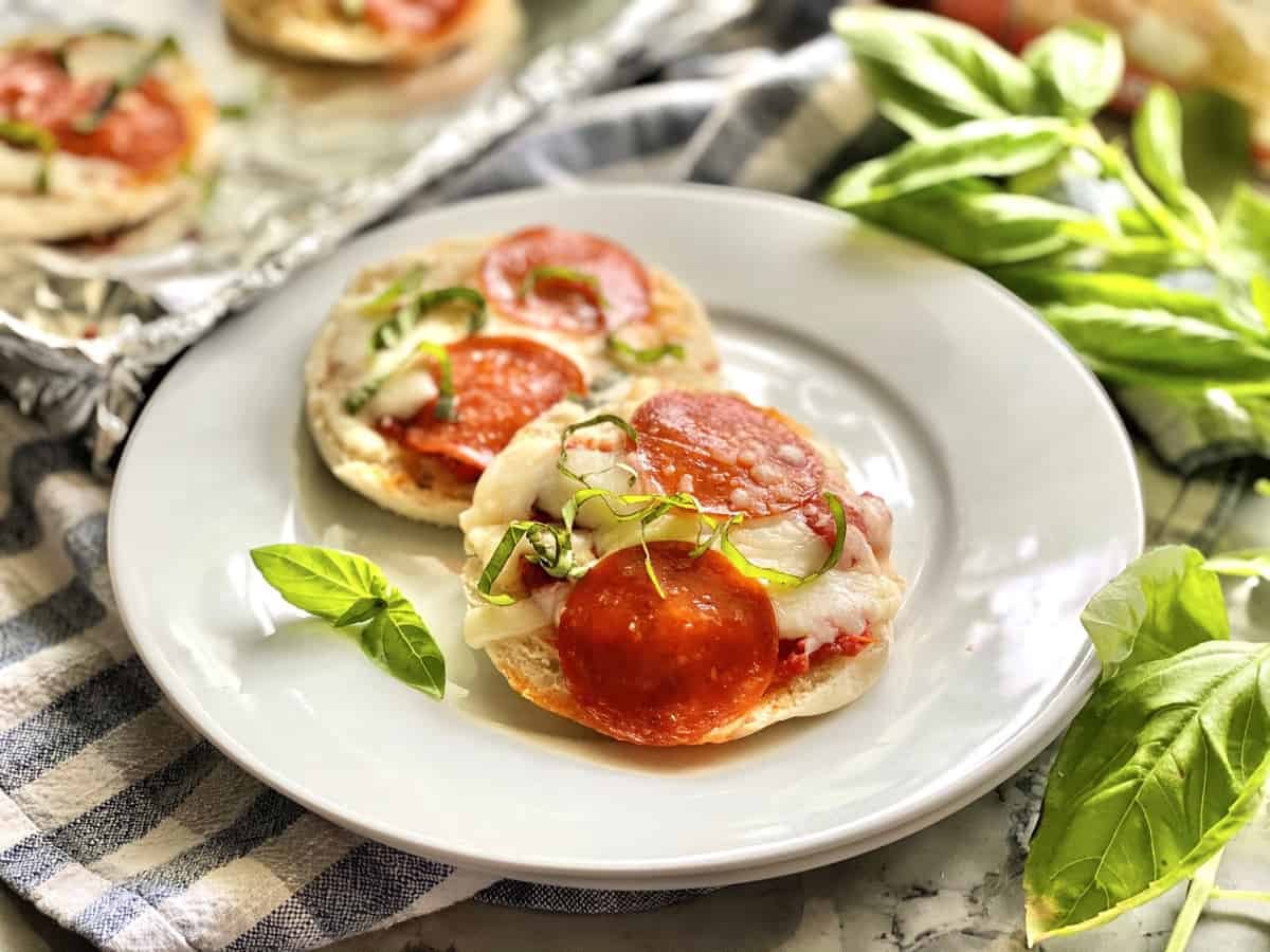 Cooked English Muffin Pepperoni Pizzas on white plate garnished with basil.