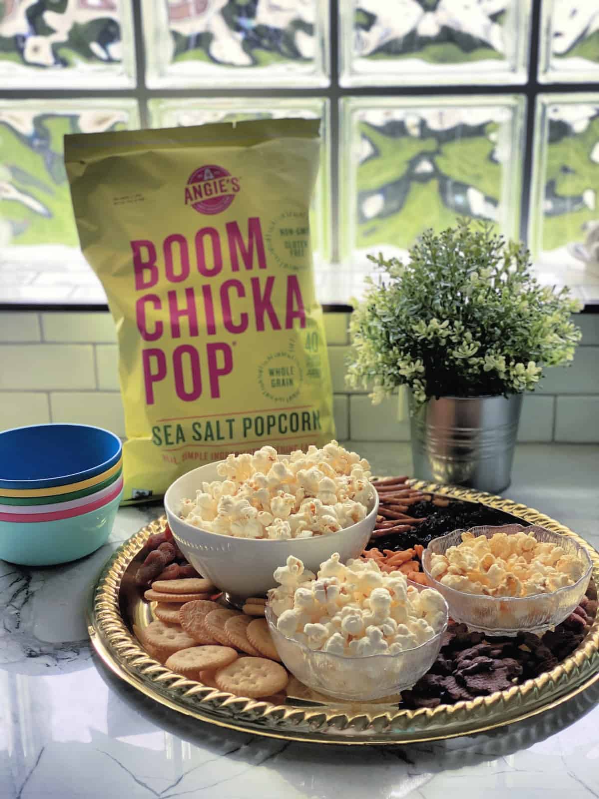 Platter with crackers, pretzels, dried fruits, and 3 bowls of popcorn in front of bag of boom chicka pop.