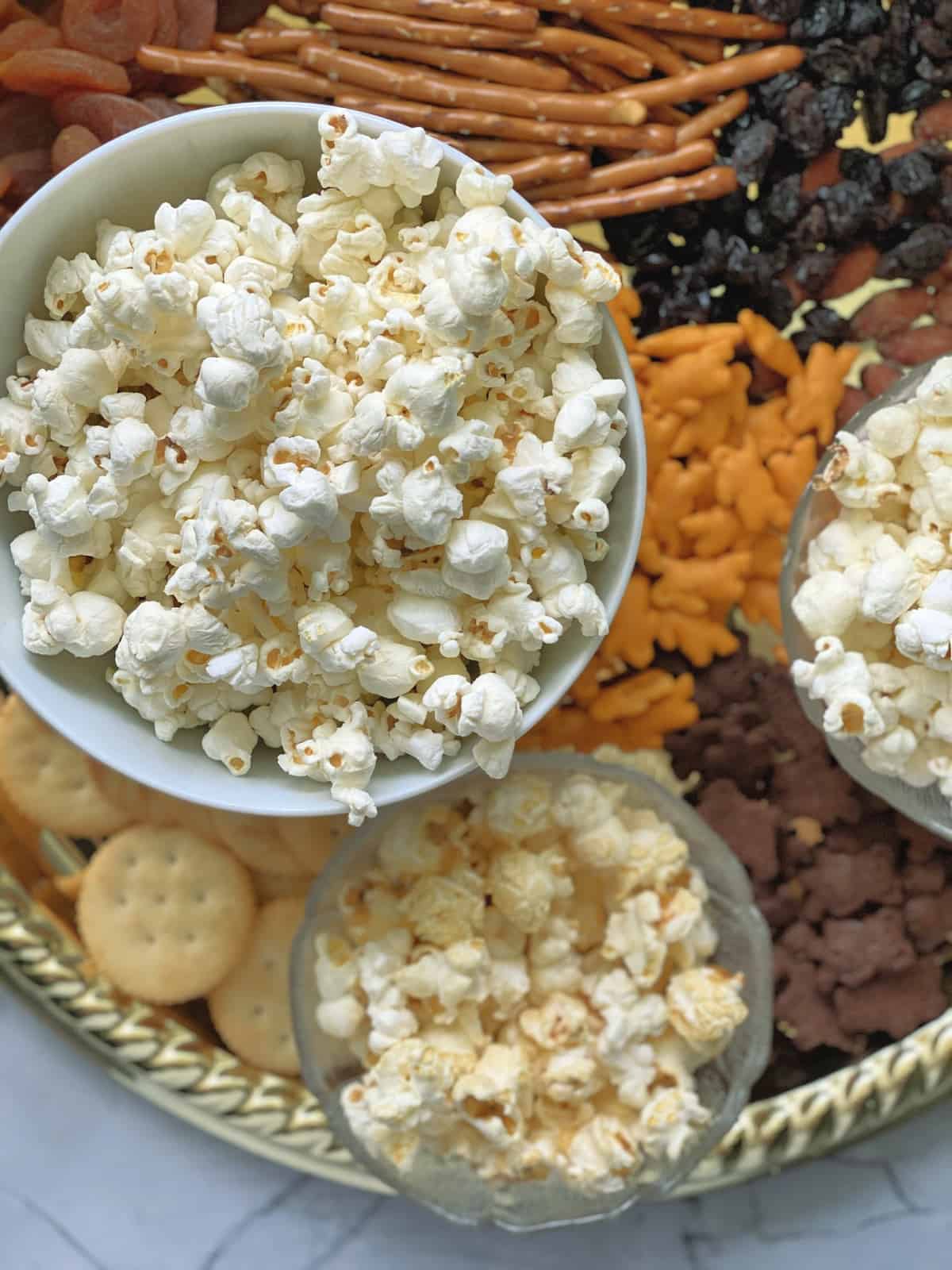 Close up top view of bowl of popcorn with pretzels, crackers, and dried fruits blurred on platter below.