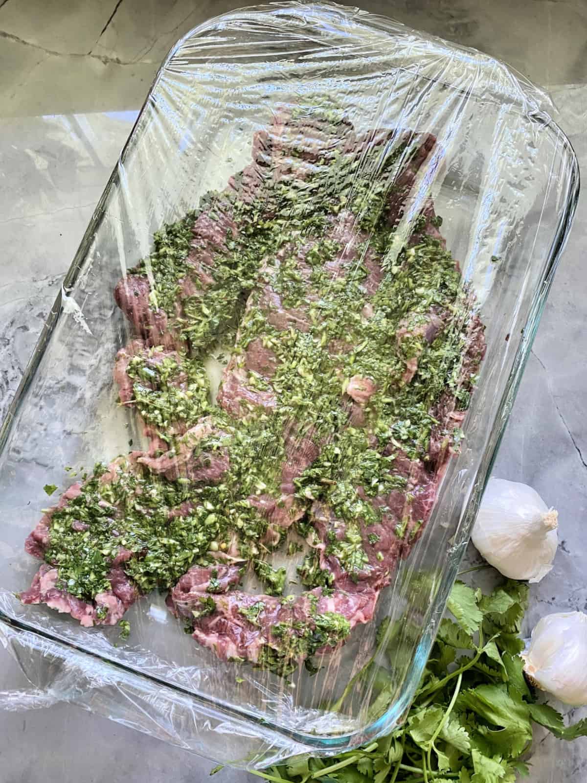 top view Grilled Skirt Steak with Chimichurri Sauce marinating in glass dish with saran wrap over top.