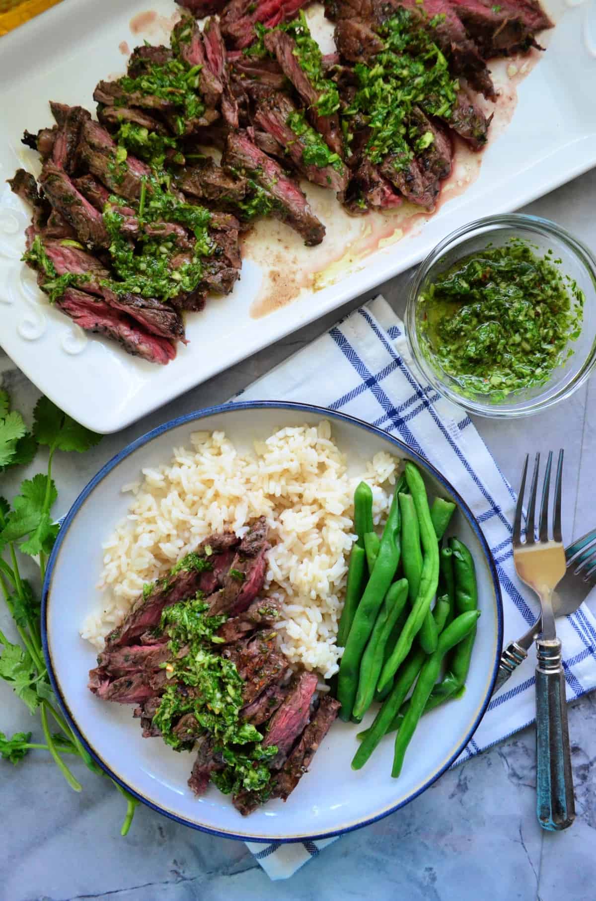 plated chimichurri sauce on grilled skirt steak with green beans over rice bed next to steak platter.