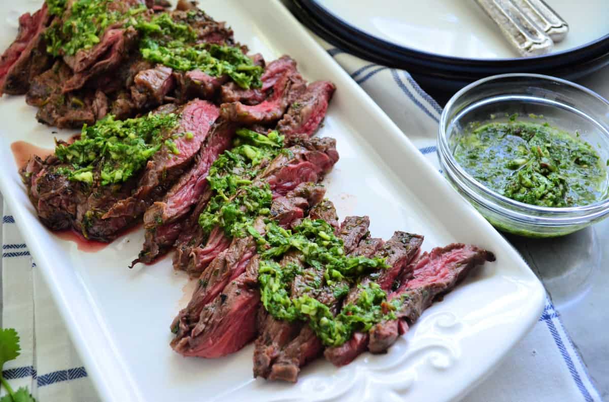 Sliced Grilled Sliced Skirt Steak on platter with Chimichurri Sauce on top and in small bowl next to it.