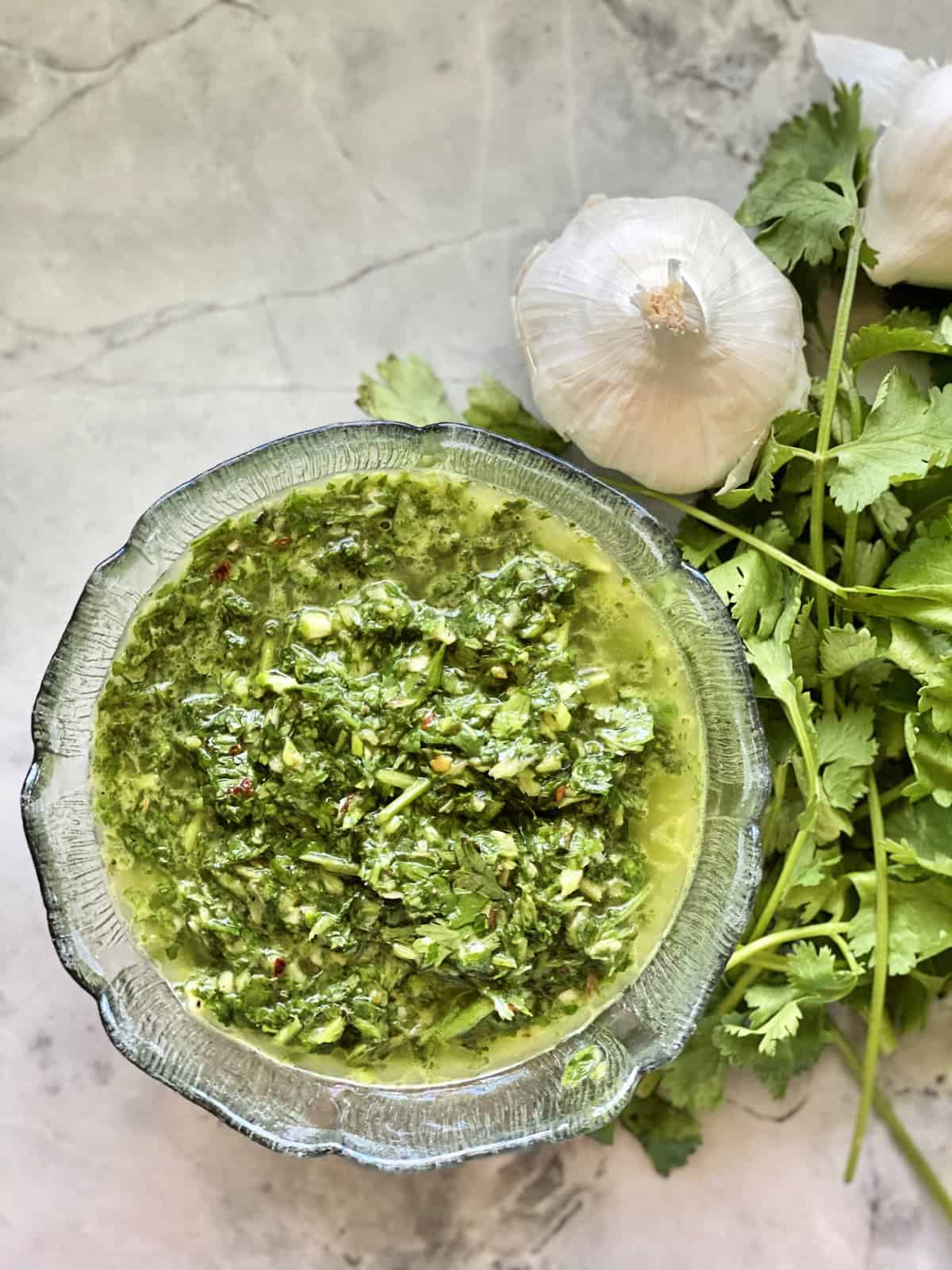 Top view of green chopped herbs and oil in glass bowl next to garlic and cilantro on counter.