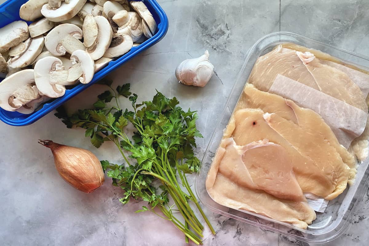 open package of chicken, bunch of parsley, garlic, sliced mushrooms, and shallot on countertop.
