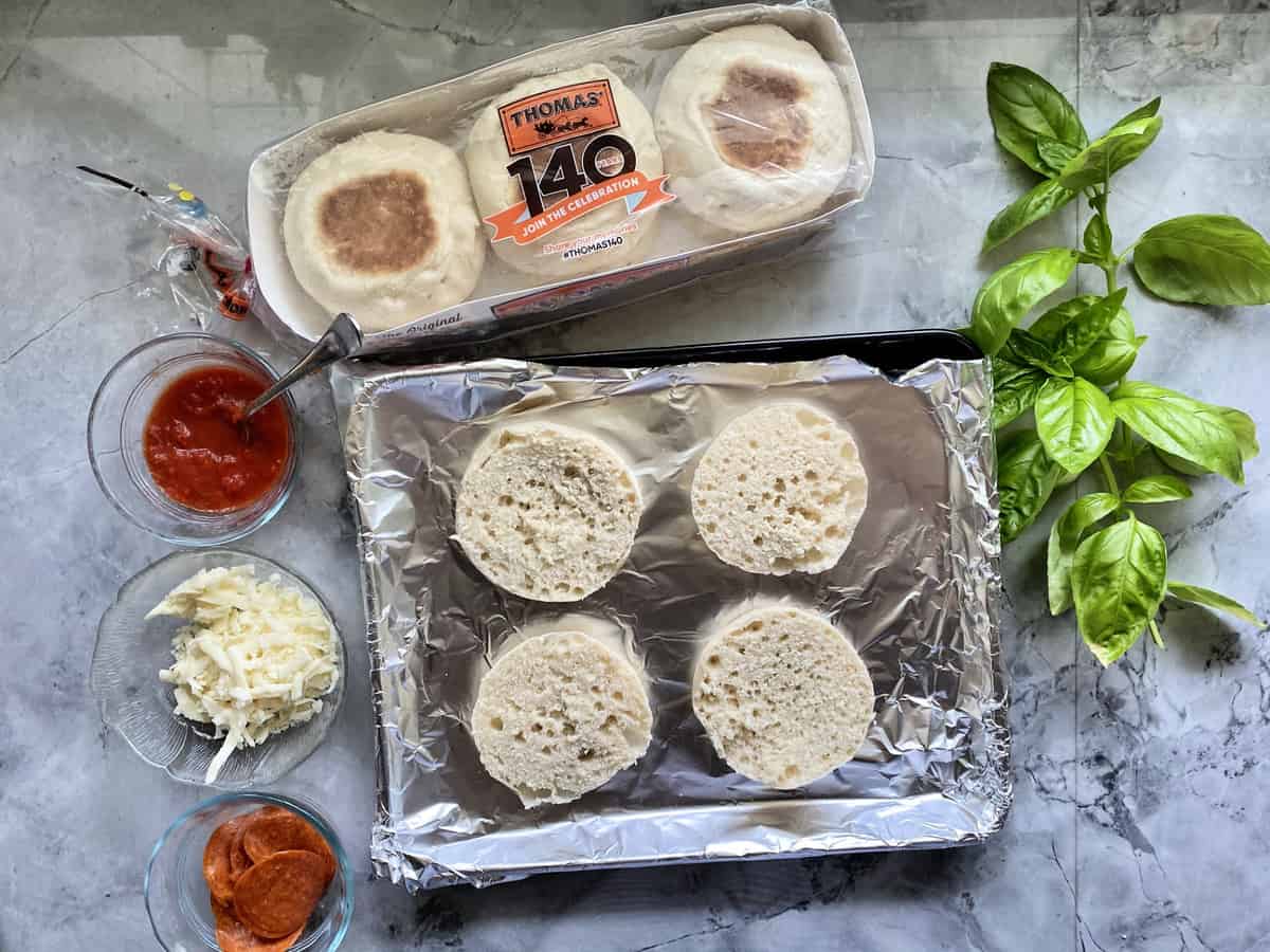 Sliced Thomas English Muffins on aluminum foil covered pan by sauce, cheese, basil, and pepperoni.