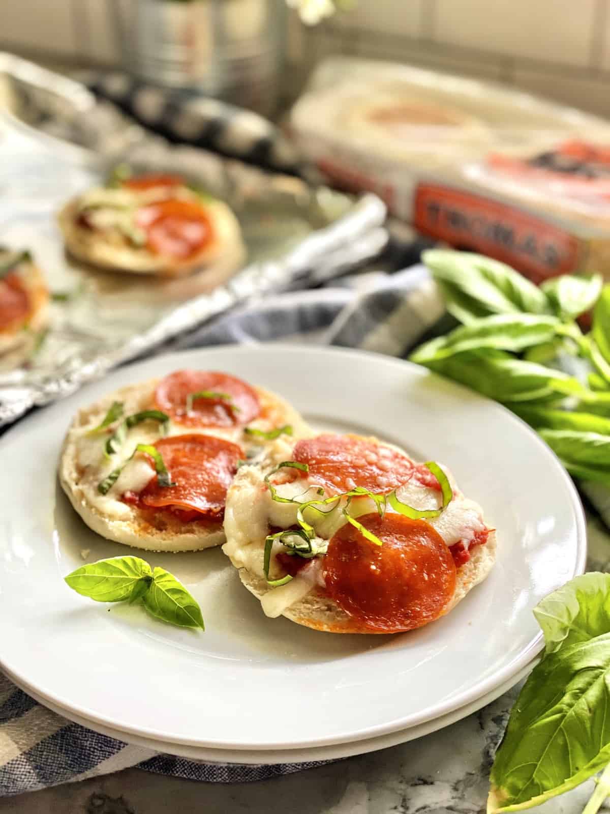 Two Toaster Oven English Muffin Pizzas garnished with basil on a white plate over checkered tablecloth.