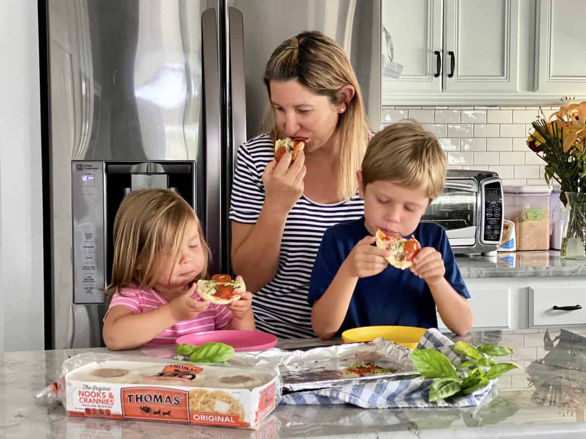 Mother and 2 children eating toaster oven english muffin pizzas over kitchen counter.