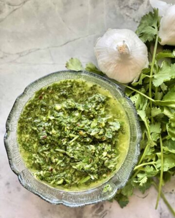 Homemade Chimichurri Sauce in glass bowl placed decoratively on countertop with garlic and cilantro.