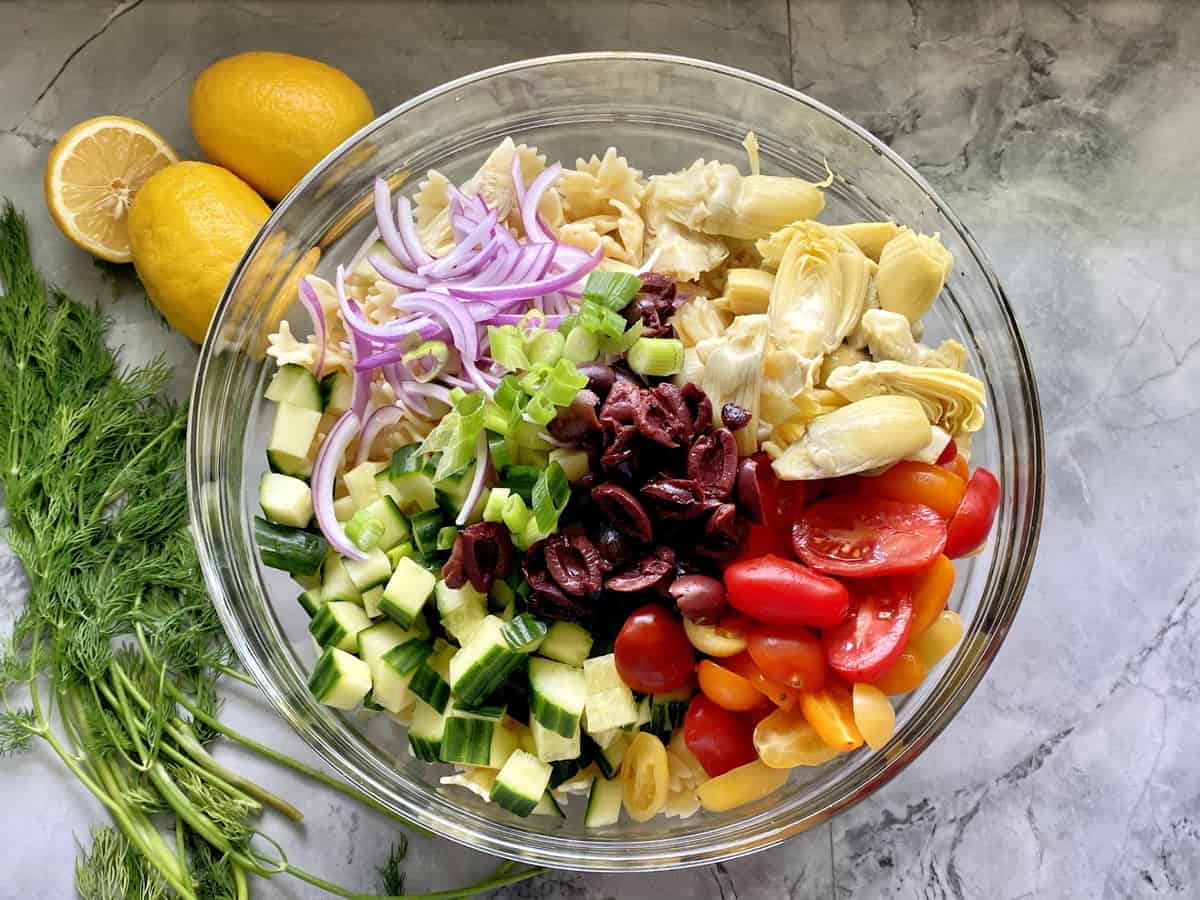 Glass bowl filled with vegetables (tomatoes, cucumbers, olives, artichoke hearts, red onions) for pasta salad.