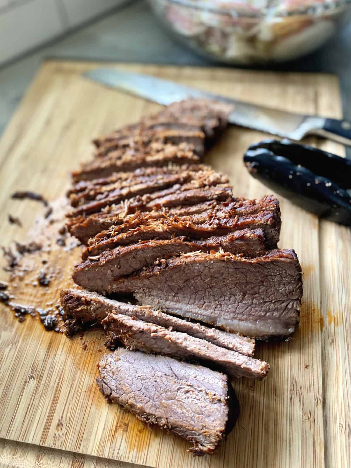 Sliced beef on a wood cutting board with a knife and tongs in the background.