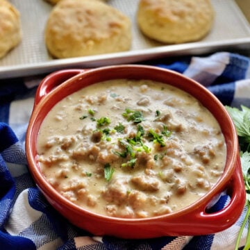 Red bowl of sausage gravy on a plaid dish cloth with biscuits in the background.