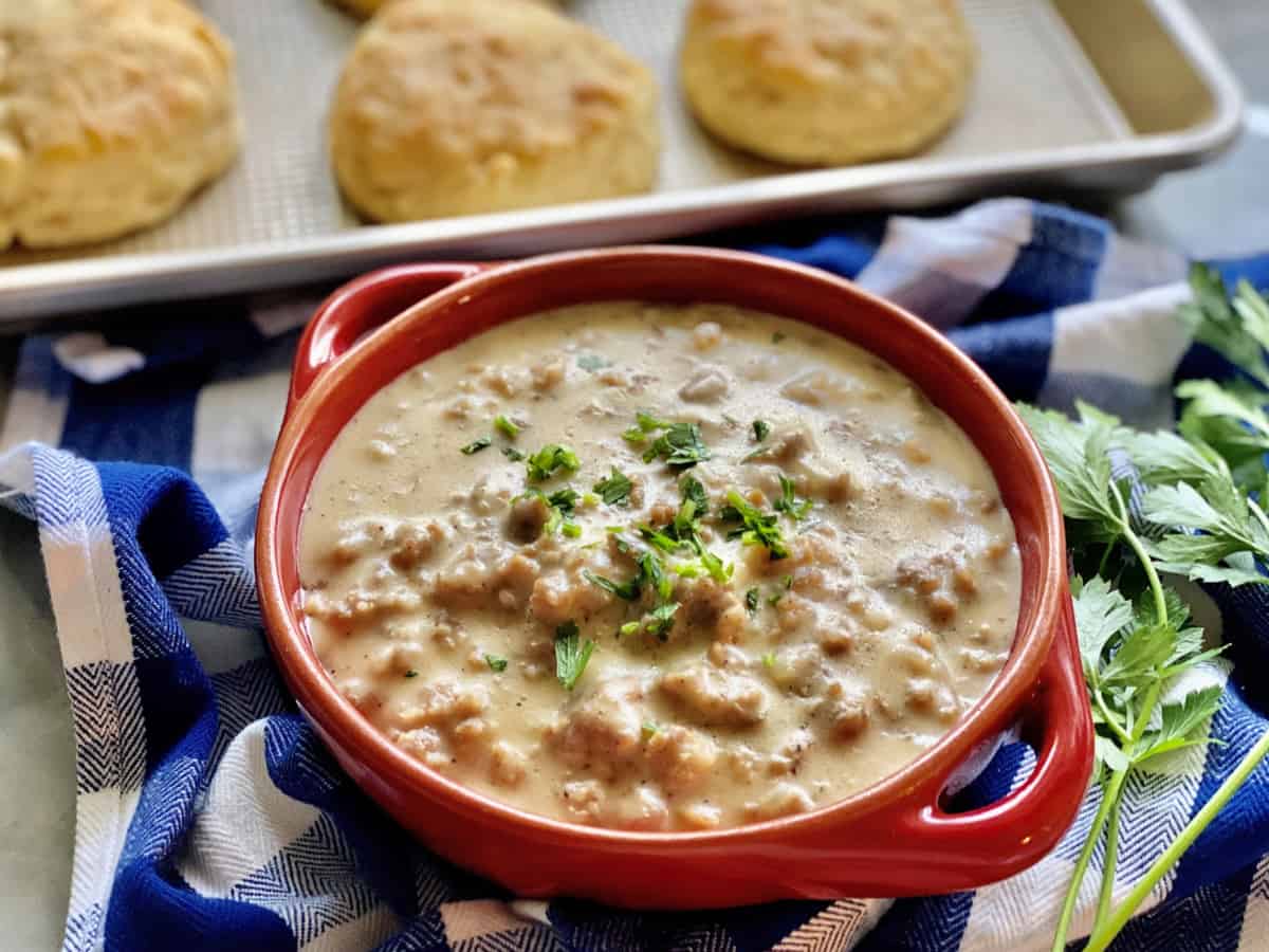 Red bowl of sausage gravy on a plaid dish cloth with biscuits in the background.