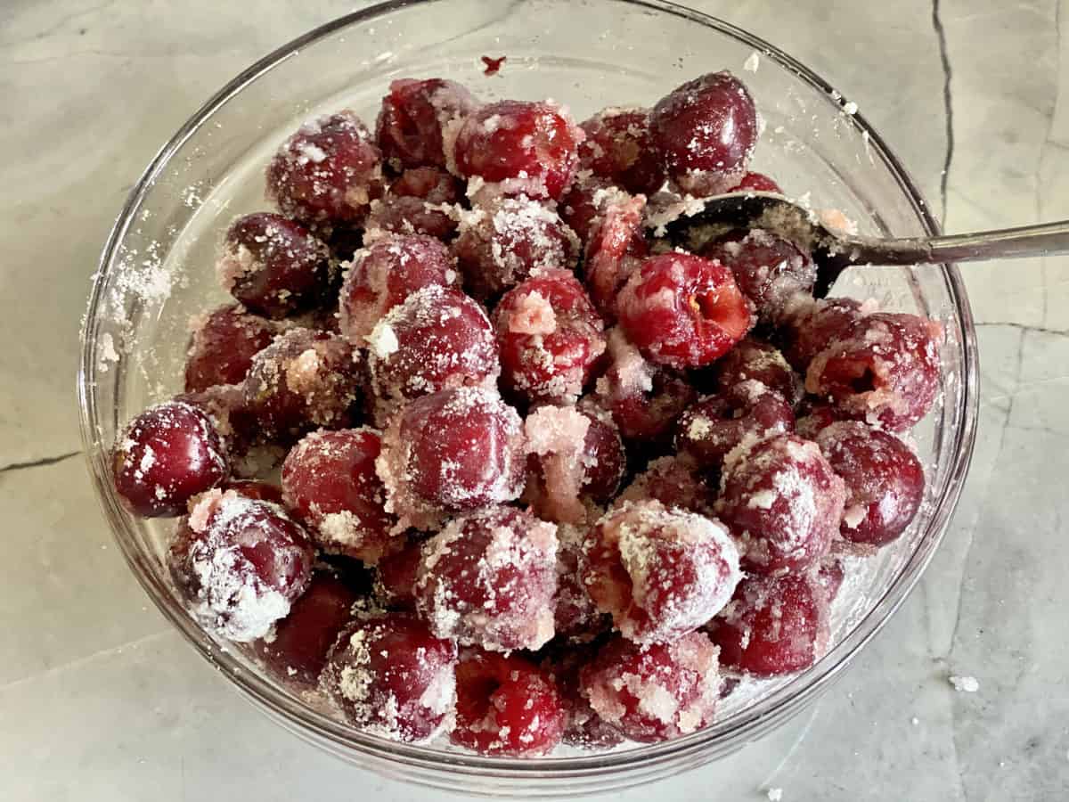 Top view of sugar coated fresh pitted cherries in a glass bowl on a marble countertop.