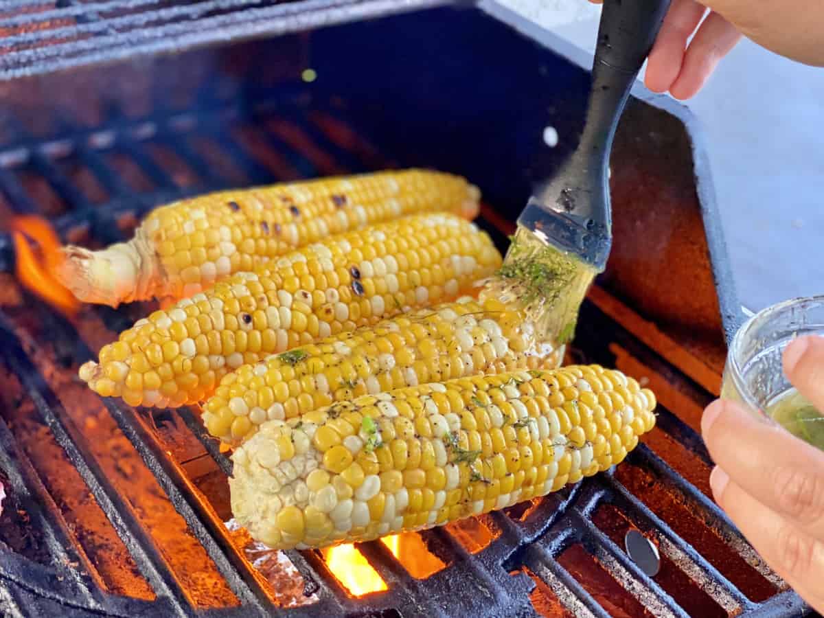 Corn on the cob on a grill with flame being basted with butter using silicone brush.