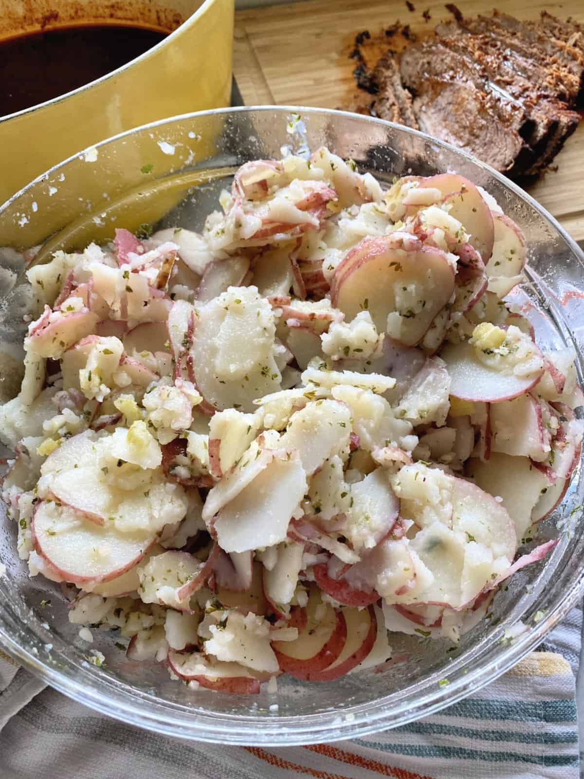 Sliced potatoes with dressing in a glass bowl with brisket in the background.