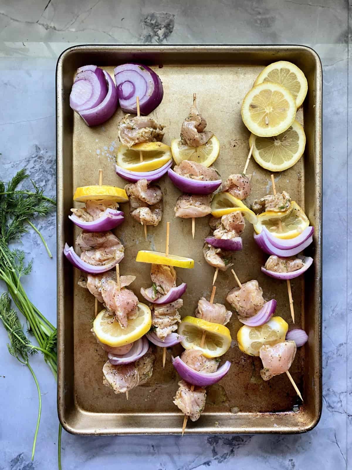 Raw chicken kabobs on a baking sheet with fresh lemon and red onion and dill.