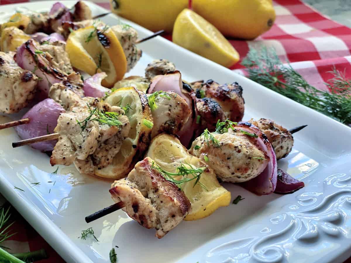 Chicken kebab with grilled onion and lemon with fresh dill on a white platter.