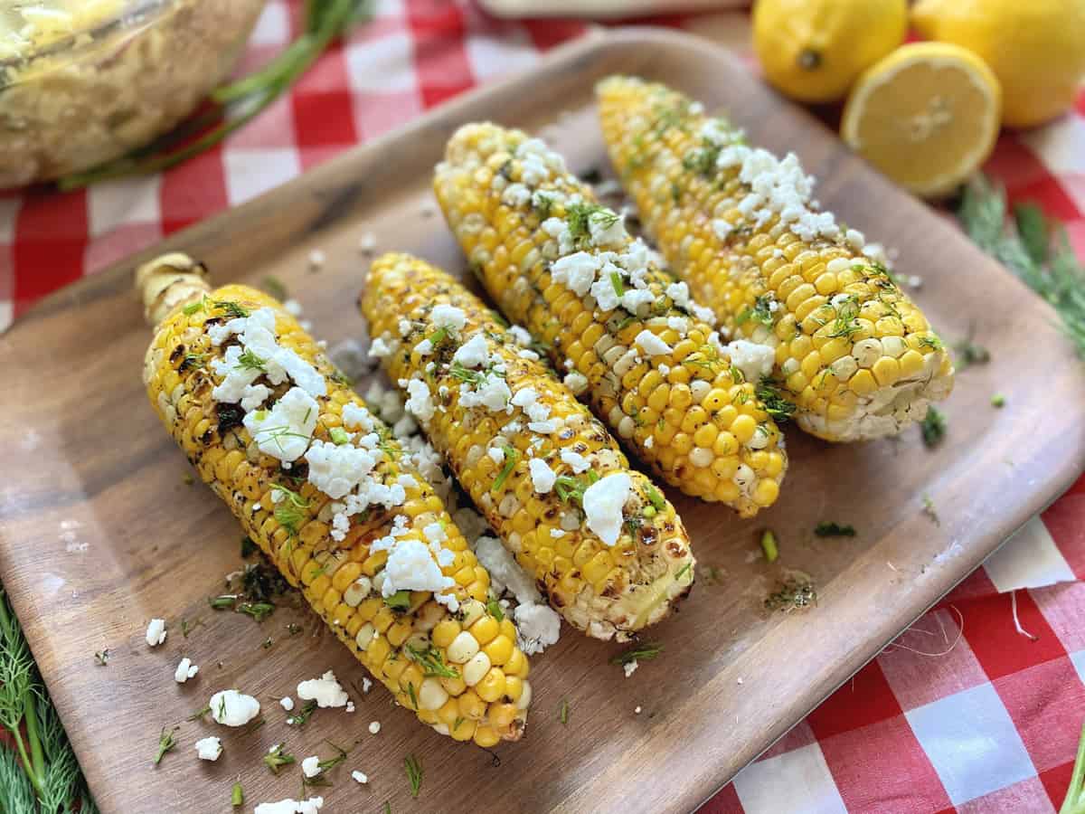 Grilled corn on the cob topped with feta and dill on a wooden platter on a red checkered table cloth.