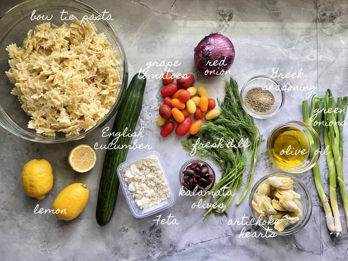 Ingredients for pasta salad, bow tie pasta, tomatoes, cucumber, lemon, feta, olive oil, red onion, and more.