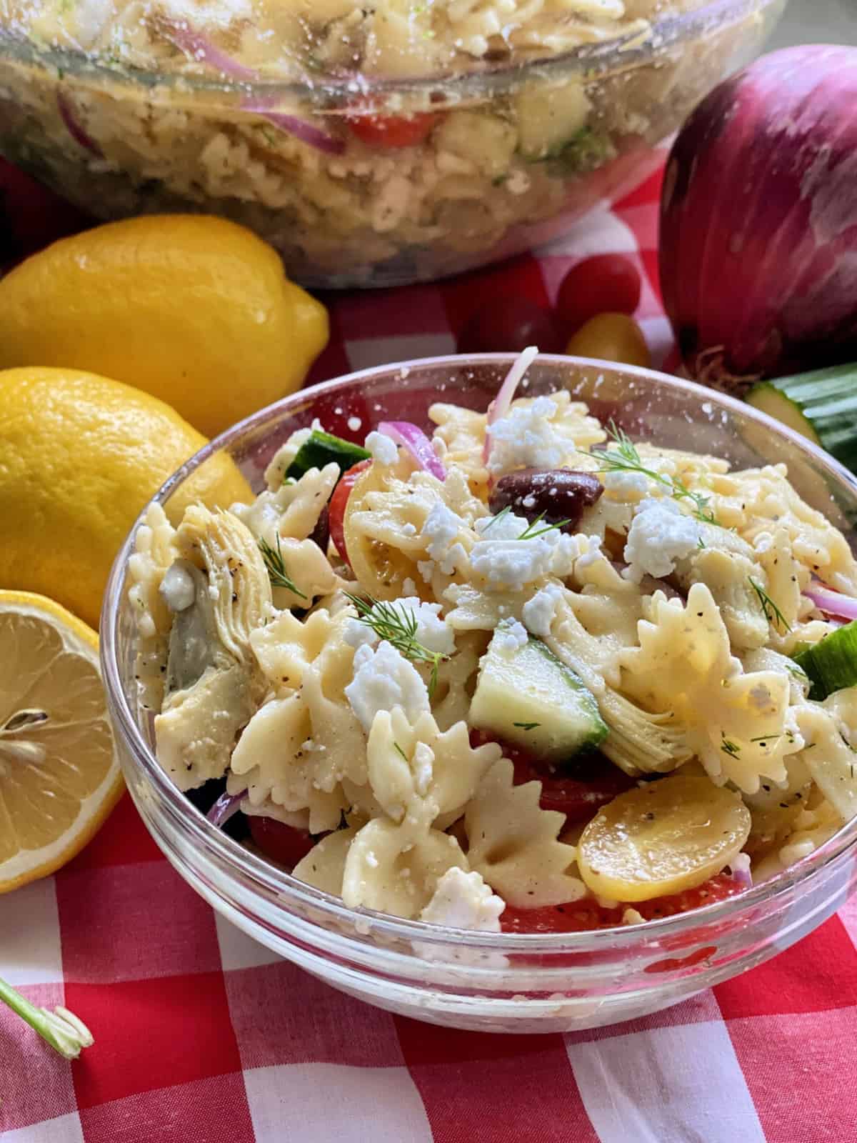 A glass bowl of pasta salad on a red and white checkered tatblecloth.