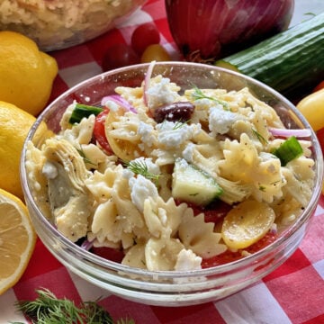 A glass bowl of bow tie pasta salad on a red and white checkered tablecloth.